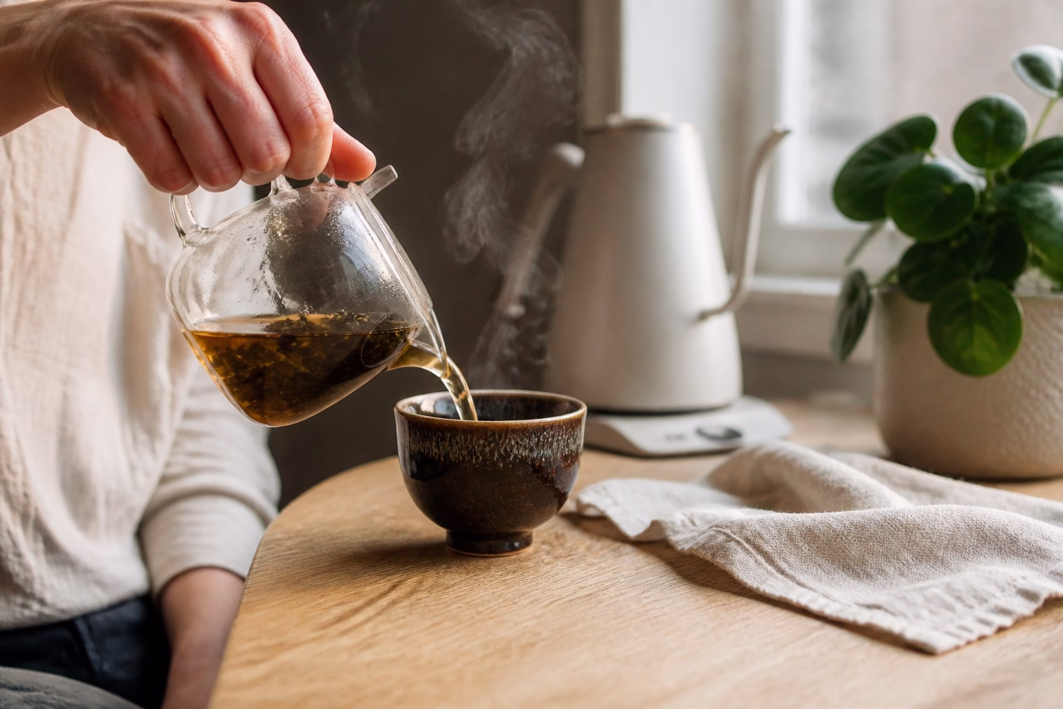 Loose leaf tea being poured into a handmade dark-glazed Jianzhan cup in a sunlit home