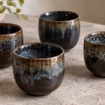 Four ceramic cups with varied dark brown and blue reactive glaze patterns on a stone table